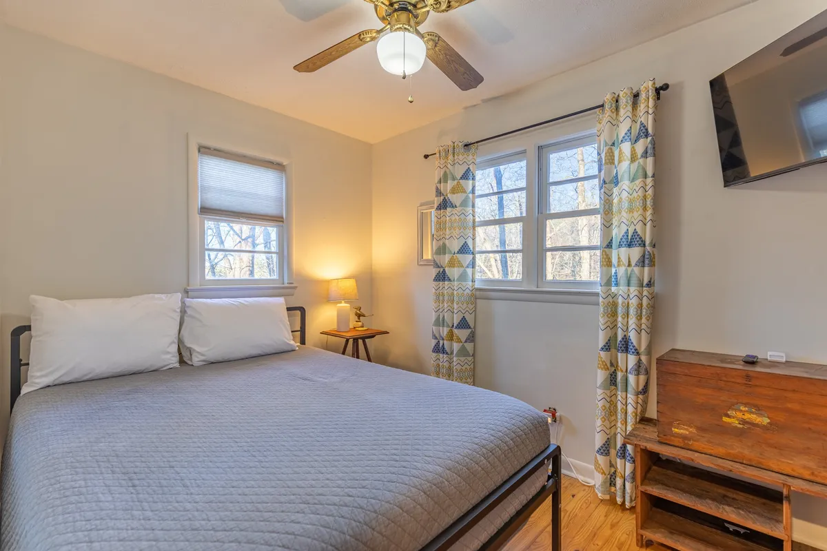 A peaceful bedroom scene with a comfortable bed, windows draped with curtains, a lamp, a TV, and a wooden chest. The room is bathed in gentle light, creating a serene atmosphere ideal for winding down or enjoying a morning coffee in bed.
