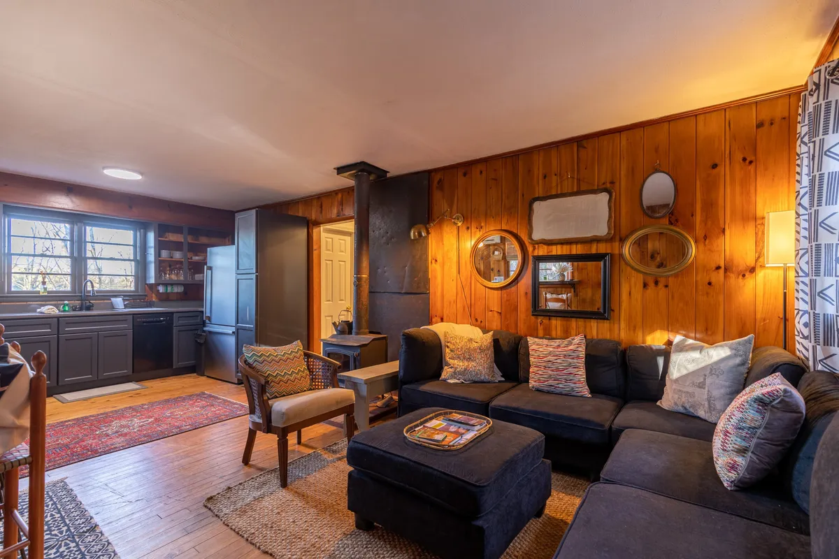 The image showcases a warm, inviting living area with a plush dark blue sectional sofa, a rustic wooden accent wall adorned with mirrors, a cozy woodburning stove, and a dining table—all bathed in natural light, creating an intimate and comfortable space ideal for relaxing or gathering with loved ones.