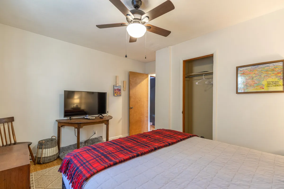This inviting bedroom features a cozy bed with a red plaid throw, a television mounted opposite the bed, and a small wooden table with a chair, offering a relaxing spot to unwind. The mood is intimate and perfect for curling up with a show or book.