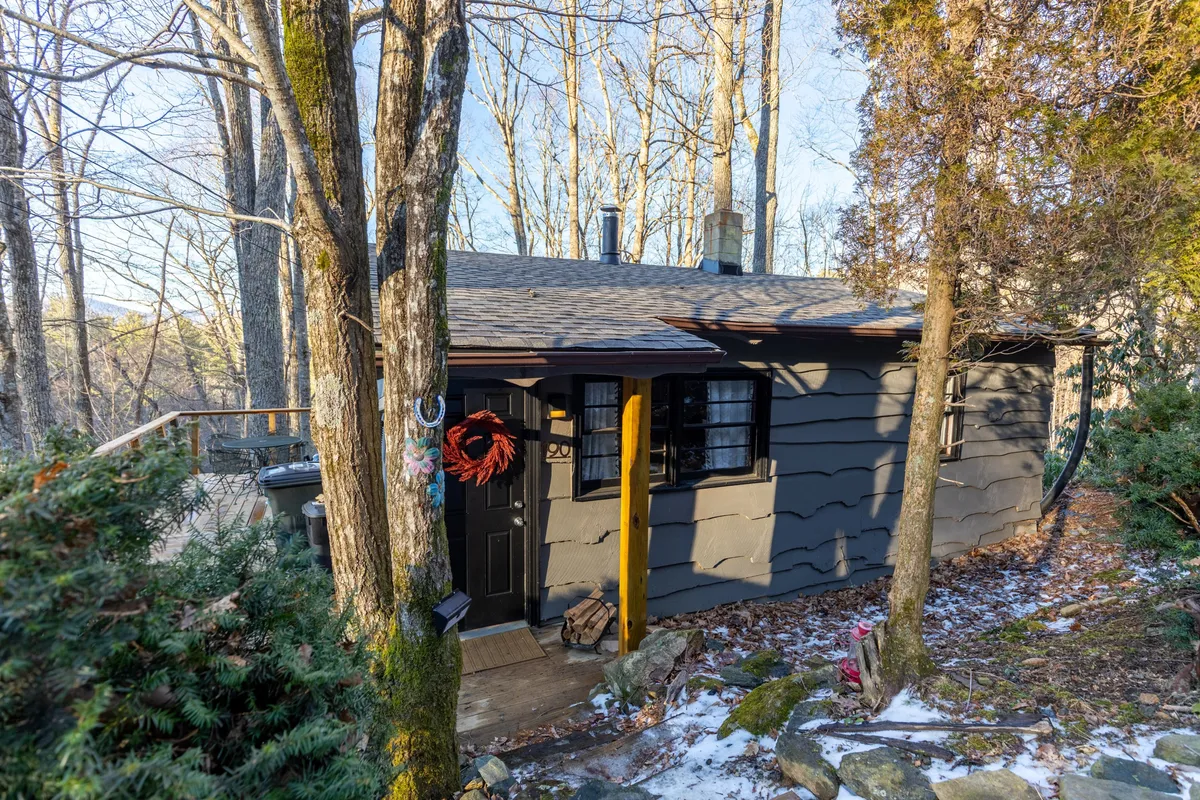 A welcoming exterior view of the cottage’s front entrance, featuring a charming wreath on the door, natural stone path, and mature trees surrounding the home. The scene hints at a peaceful, nature-rich neighborhood ideal for strolls and outdoor relaxation.