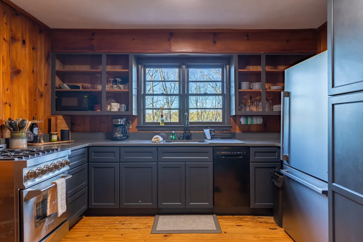 A cozy, updated kitchen with a gas stove as the centerpiece, accented by open shelving and large windows that let in natural light. Black appliances add a modern touch, and the inviting setup is ideal for preparing meals or sharing a morning coffee while overlooking the scenic outdoors.