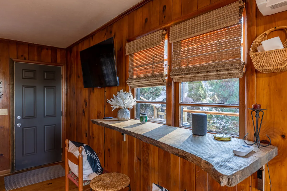 This scene highlights the breakfast bar with wooden counter, bar stools, nearby windows, decorative plants, and a television visible in the background. The setup encourages casual snacking, planning, and socializing in a bright and cozy nook.