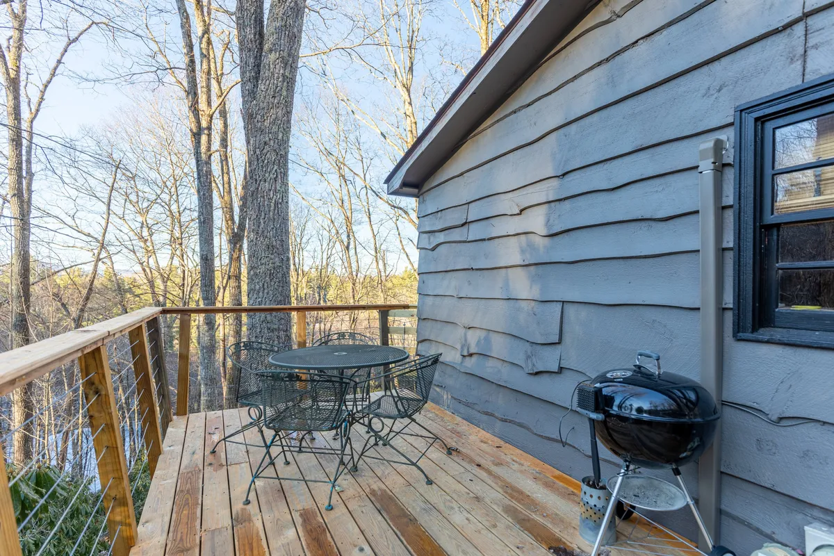 The image shows a tranquil wooden deck set amidst lush trees, furnished with a sturdy metal patio table and chairs. A charcoal grill sits close by, ready for outdoor barbecues and gatherings. The setting is serene, with glimpses of the surrounding woods, suggesting a private, relaxing space perfect for dining, socializing, or simply enjoying nature.