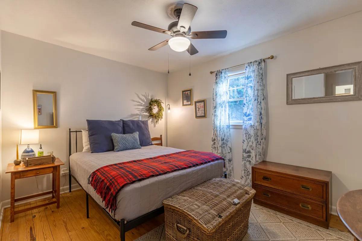 This image features a bed adorned with a red plaid blanket and decorative pillows, accompanied by a wooden nightstand with a lamp, a woven storage basket, and a window with curtains that lets in natural light. The ambiance is warm and welcoming, inviting relaxation and rest.