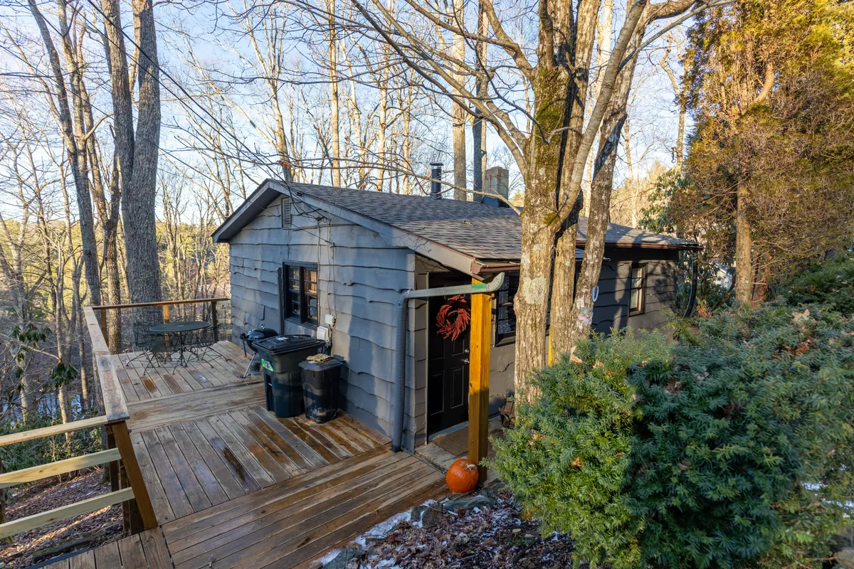 A welcoming wooden deck with comfortable outdoor furniture sits nestled among mature trees, offering a cozy atmosphere for relaxation. The deck leads to a front door, blending indoor and outdoor living. Sunshine filters through the foliage, highlighting spaces ideal for morning coffee, sunset drinks, or simply soaking up the mountain air.